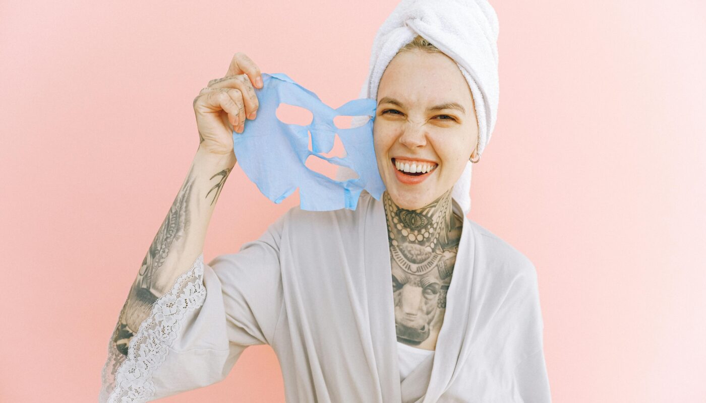 Happy woman holding a blue face mask, wrapped in a towel and smiling brightly.